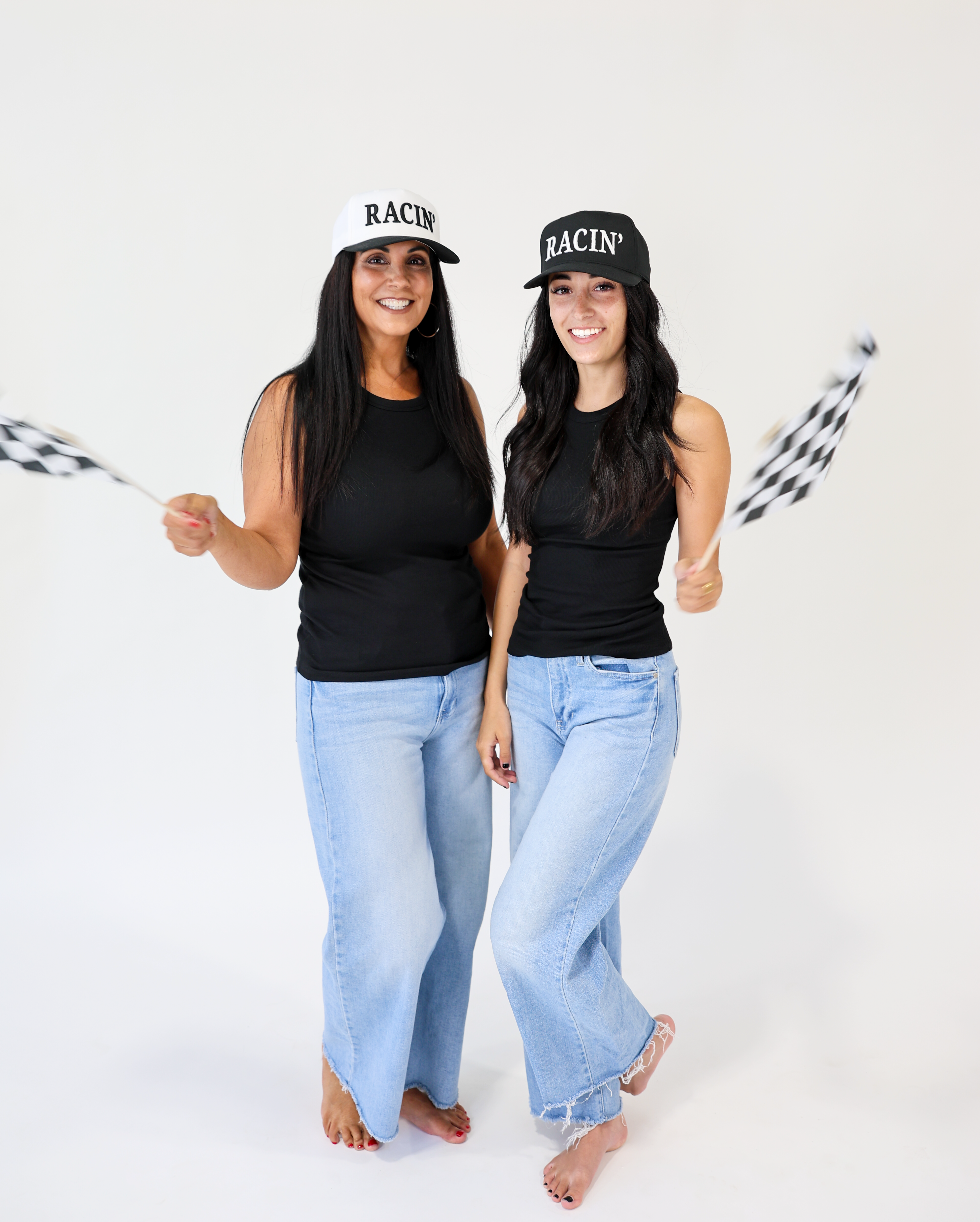 Two women wearing black tank tops and light blue jeans, holding checkered flags against a white background.
