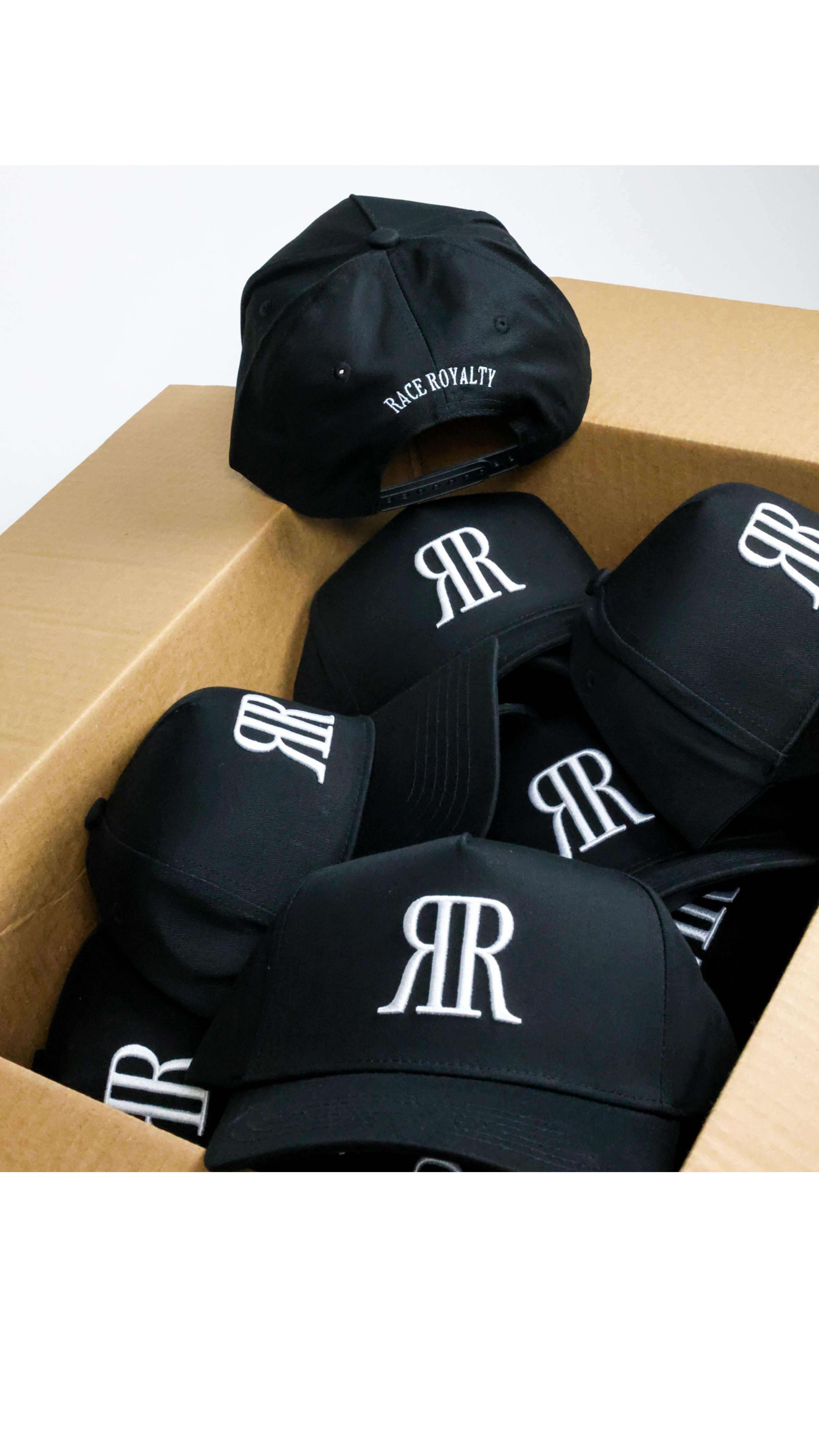 Black baseball caps with a white stylized 'RR' logo and 'RAKE ROYALTY' text on the brim, shown inside a brown cardboard box.