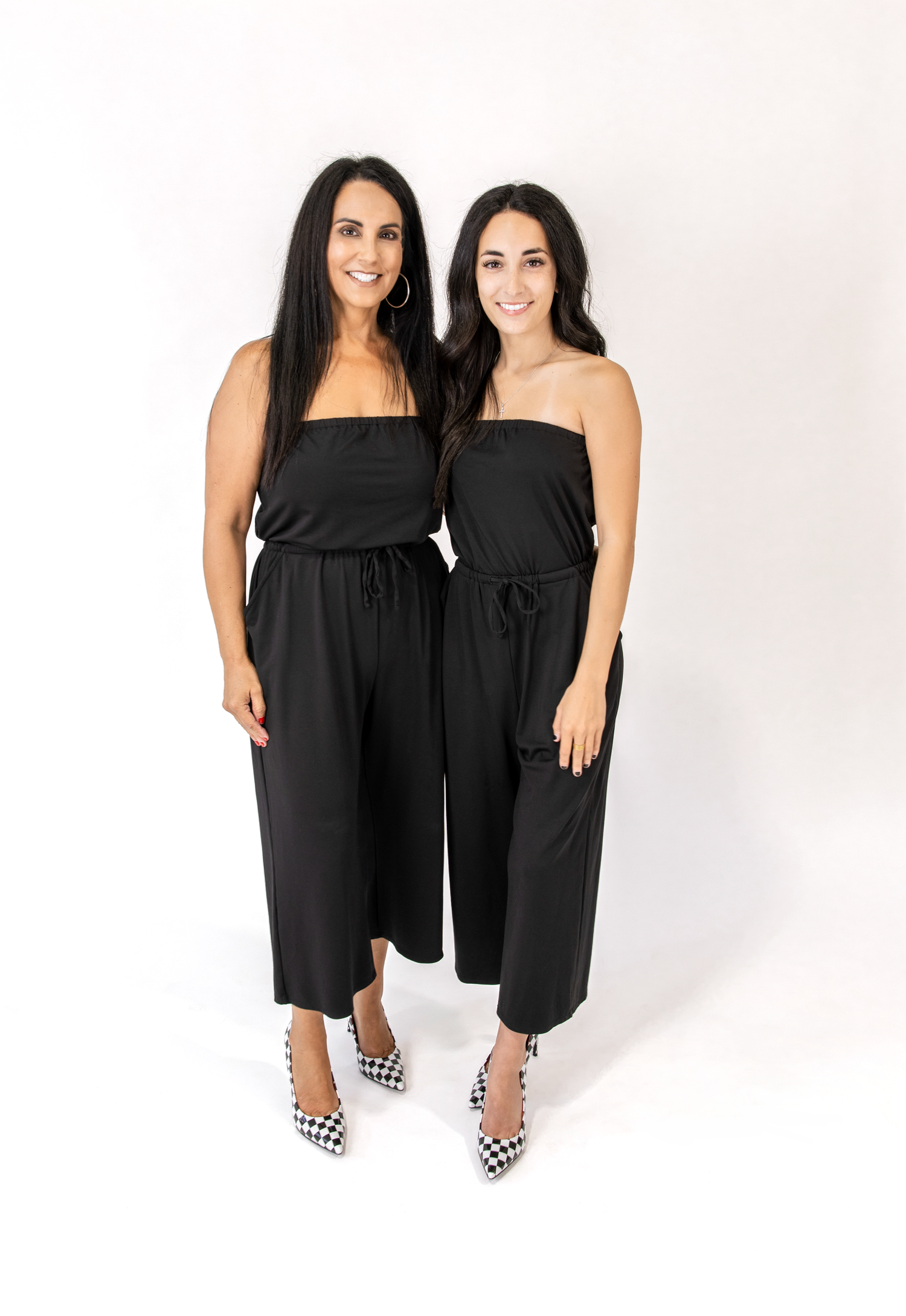 Two women standing side by side wearing matching black strapless wide-leg jumpsuits with drawstrings, paired with black-and-white checkered heels, against a plain white studio background.