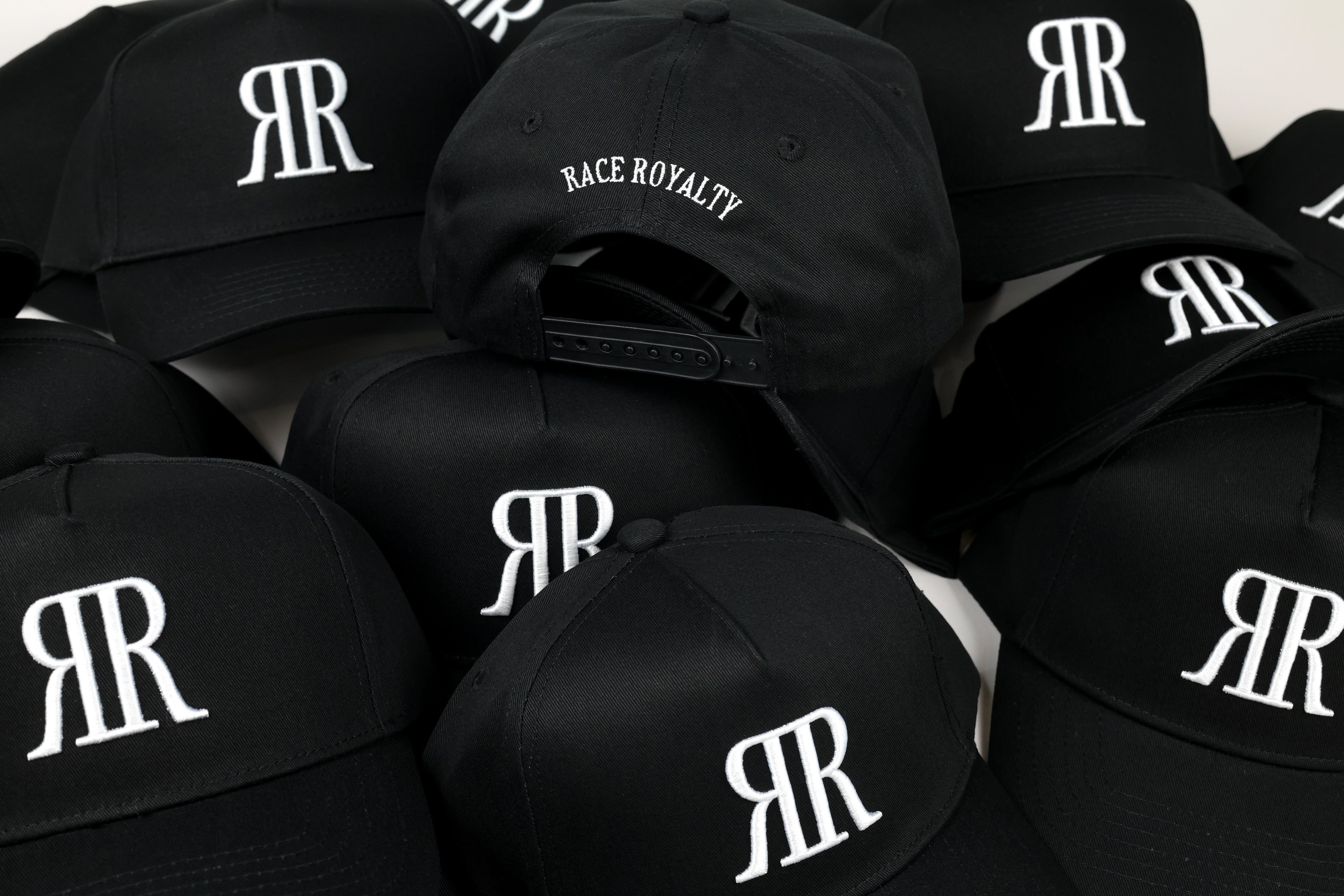 A pile of black baseball caps featuring a white embroidered 'RR' logo and one cap with 'RACE ROYALTY' text on the front, photographed against a light background.