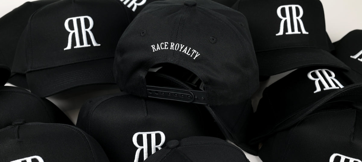 A pile of black baseball caps featuring a white embroidered 'RR' logo and one cap with 'RACE ROYALTY' text on the front, photographed against a light background.