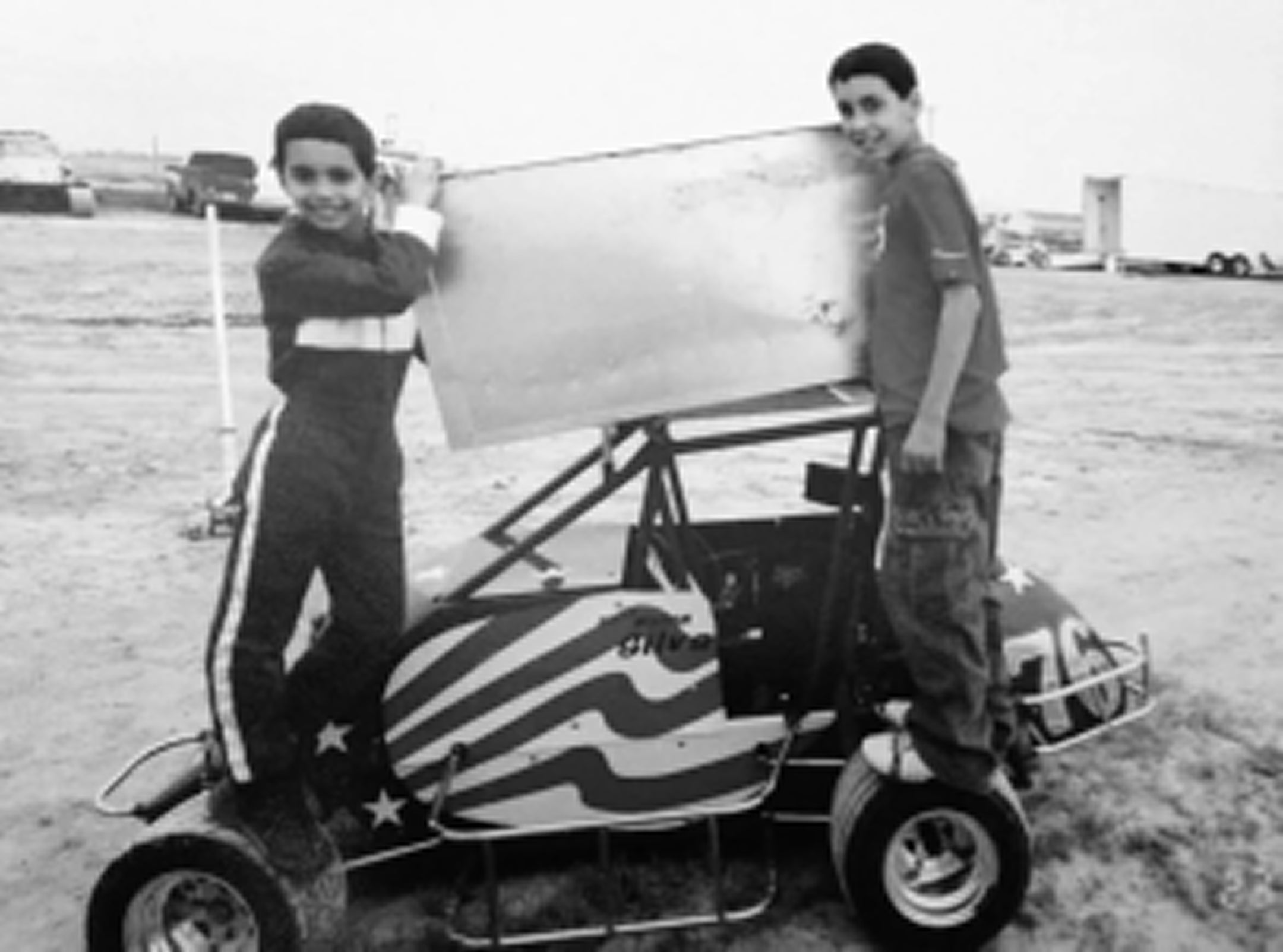 Two children standing next to a small race car with an American flag design on a grassy field.