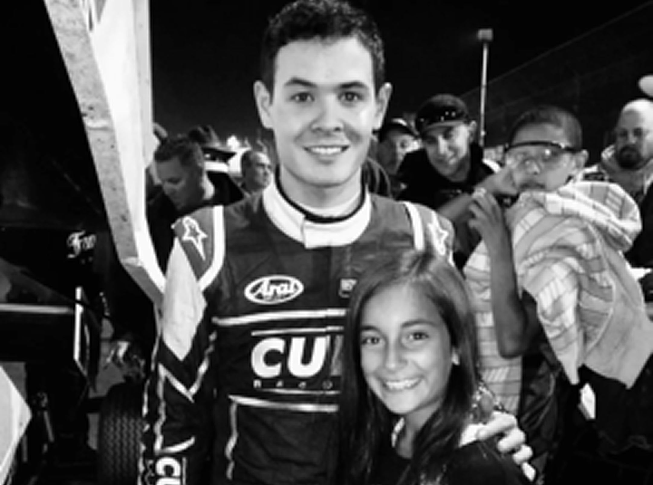 Person in racing suit with a child, surrounded by people in a casual setting
